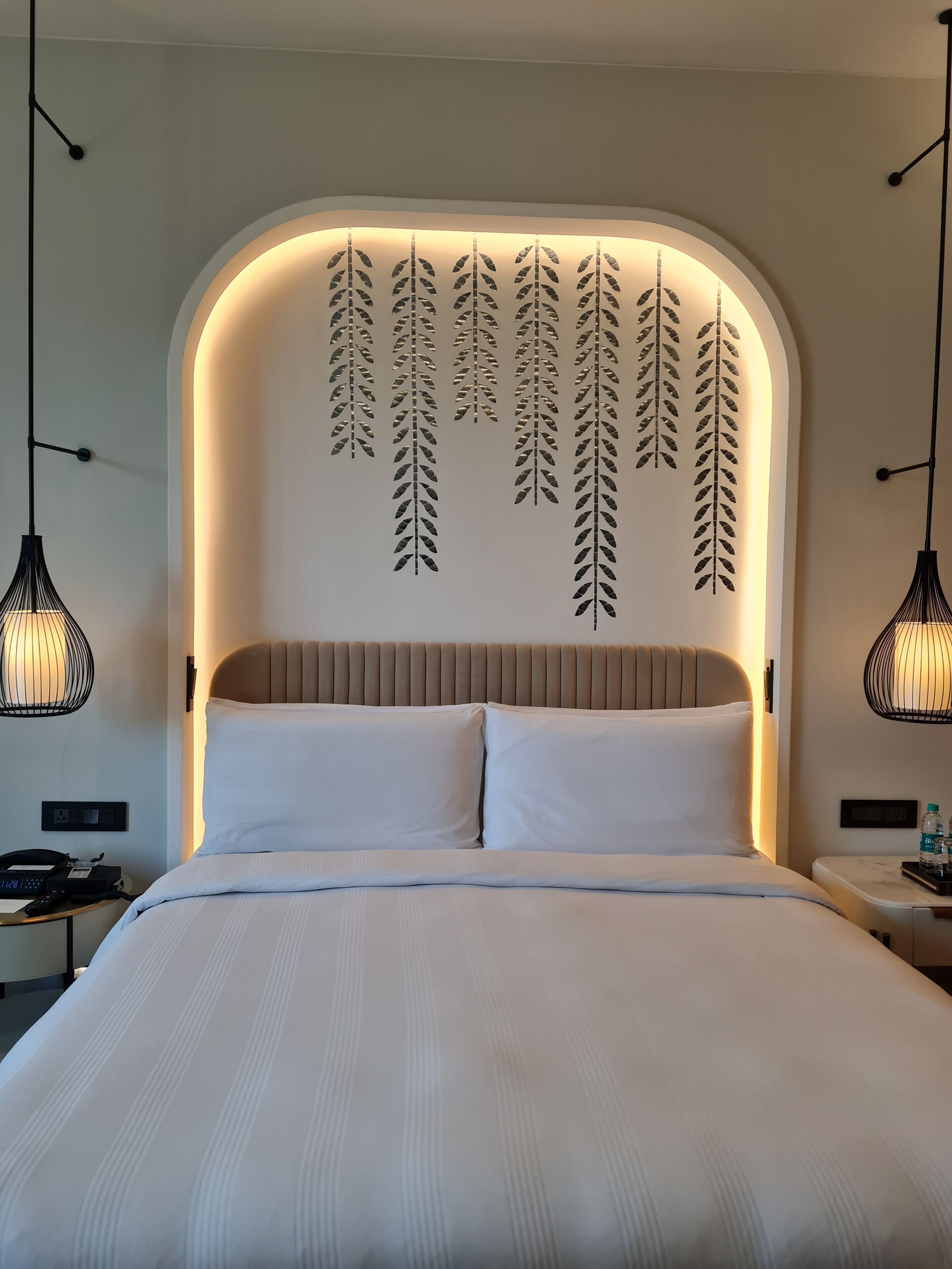 A modern hotel room with a large bed, a headboard with leaf patterns, and pendant lights.