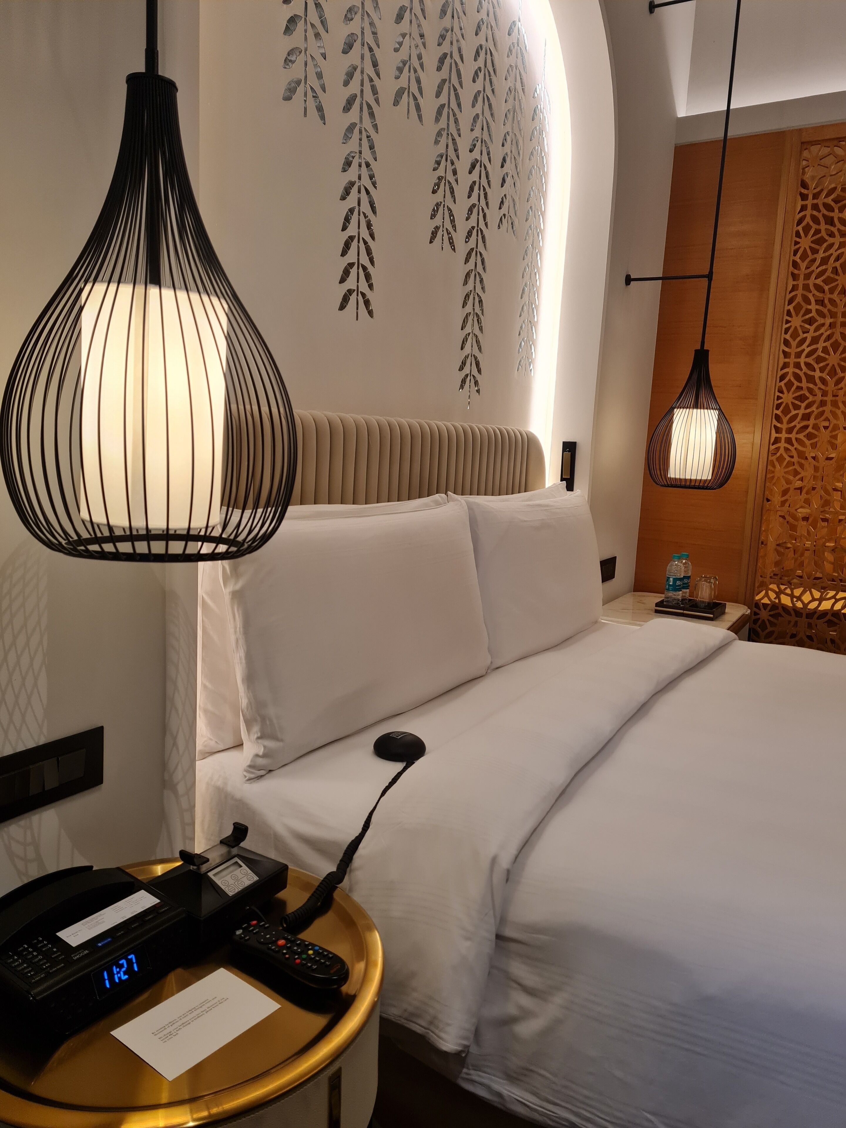 A hotel room with a bed, a bedside table with a phone and a digital clock, a hanging pendant light, and decorative wall art.