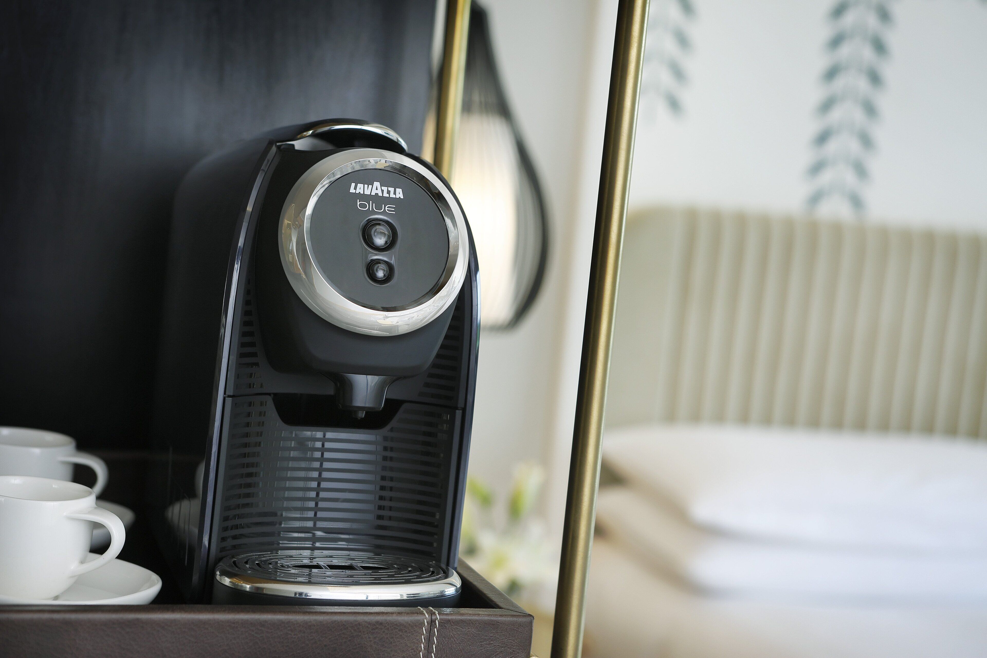 A coffee machine on a table with a cup and saucer, a bed, and a wall with a plant.