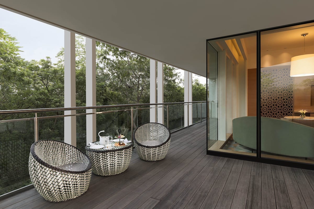 Een balkon met rieten stoelen en een tafel, grote ramen en uitzicht op bomen.