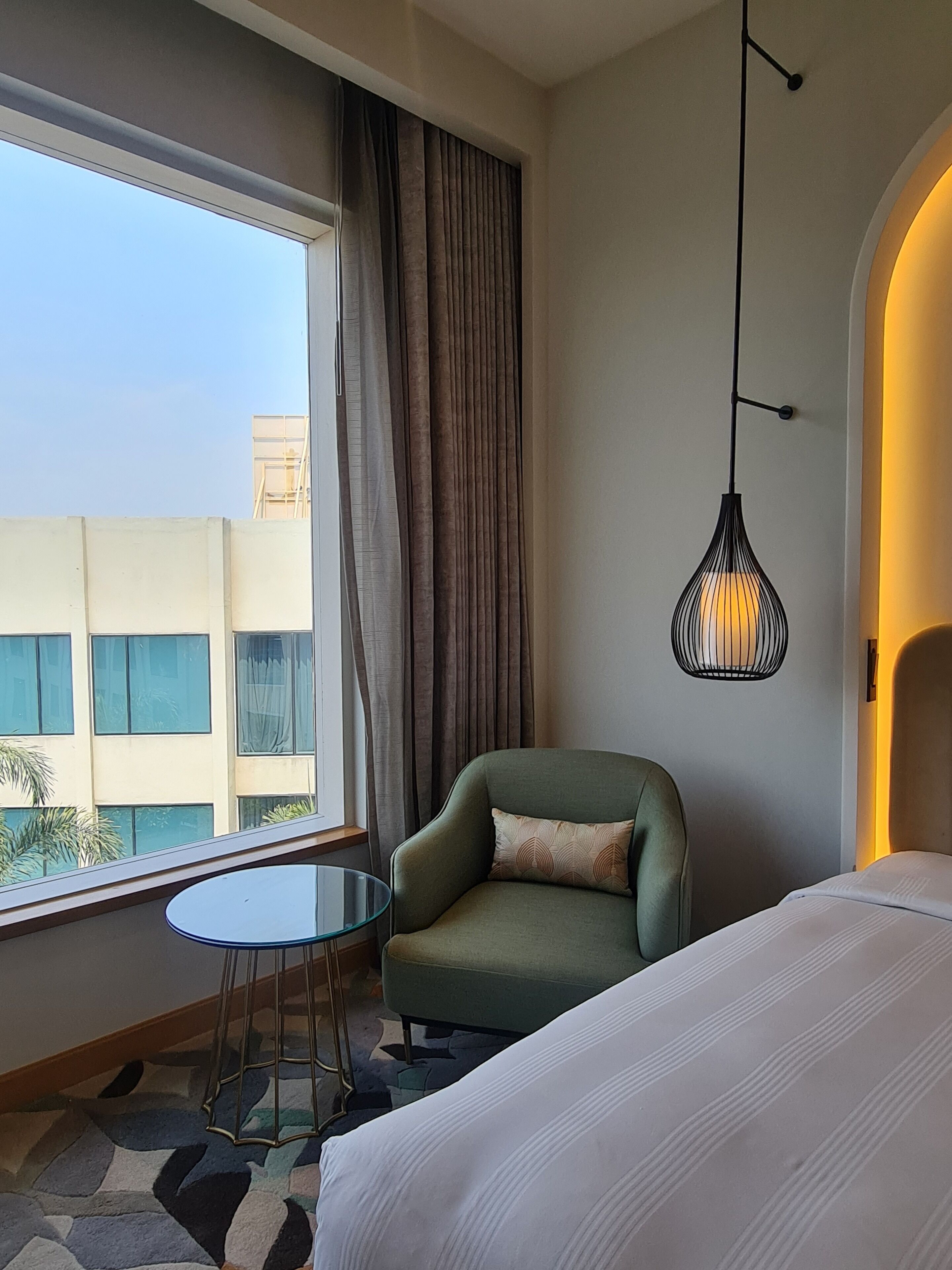 Een hotelkamer met een bed, een groene fauteuil, een klein rond tafeltje, een hanglamp en uitzicht op gebouwen.