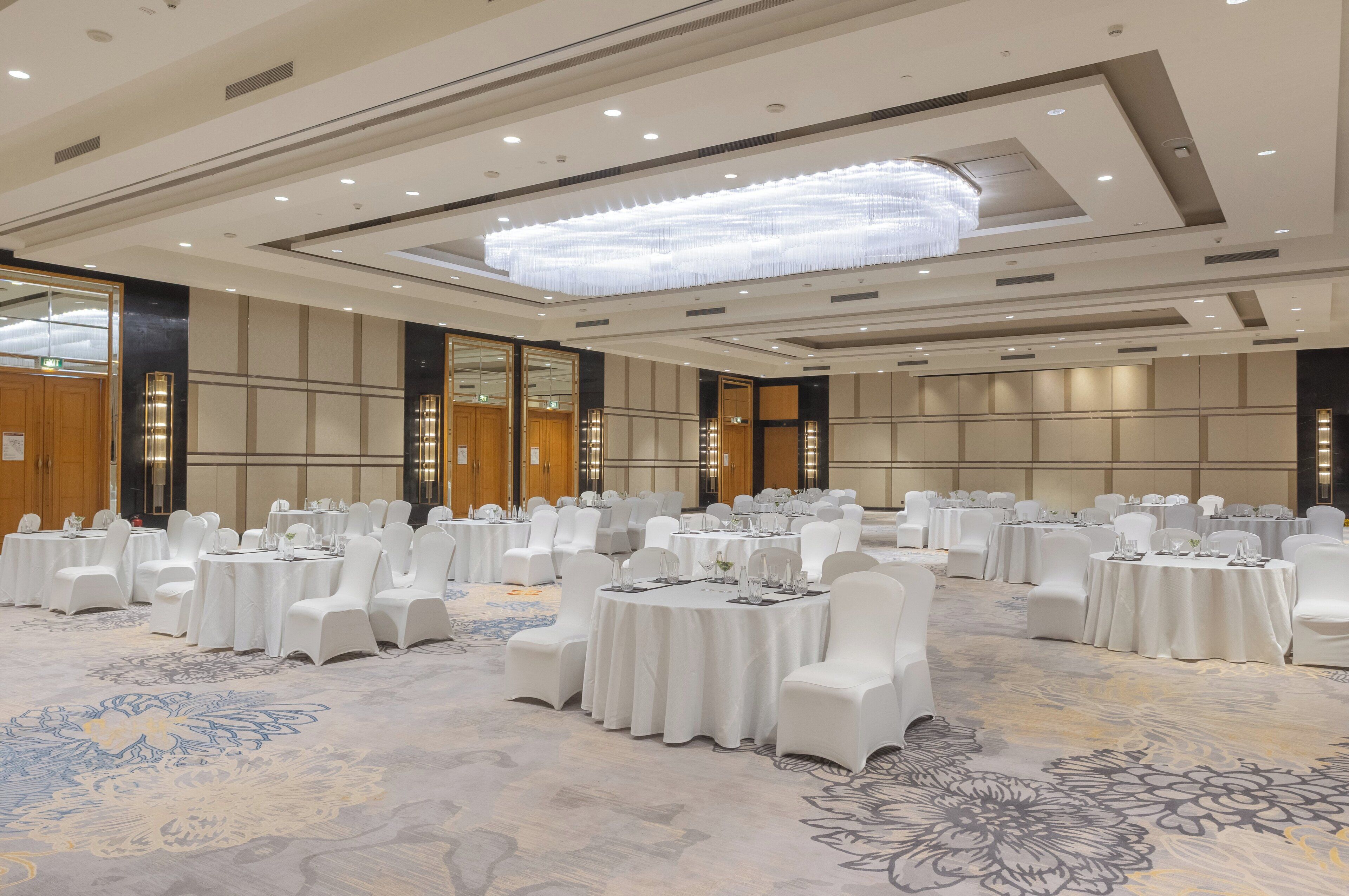 Un amplio y bien iluminado salón de banquetes con mesas redondas y sillas blancas, que incluye una araña y una alfombra estampada.
