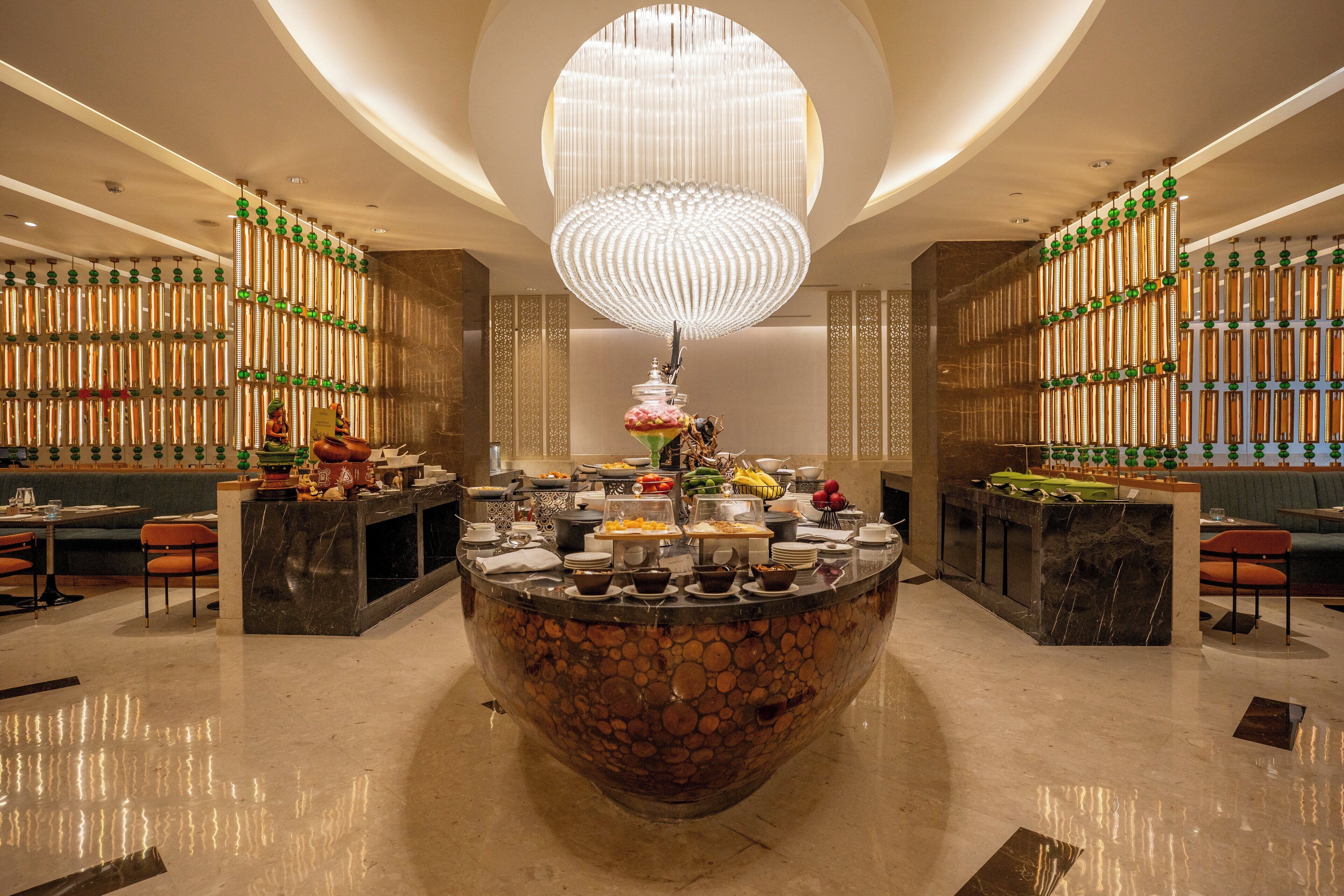 A modern hotel lobby with a central island buffet, a large chandelier, and decorative wall panels.