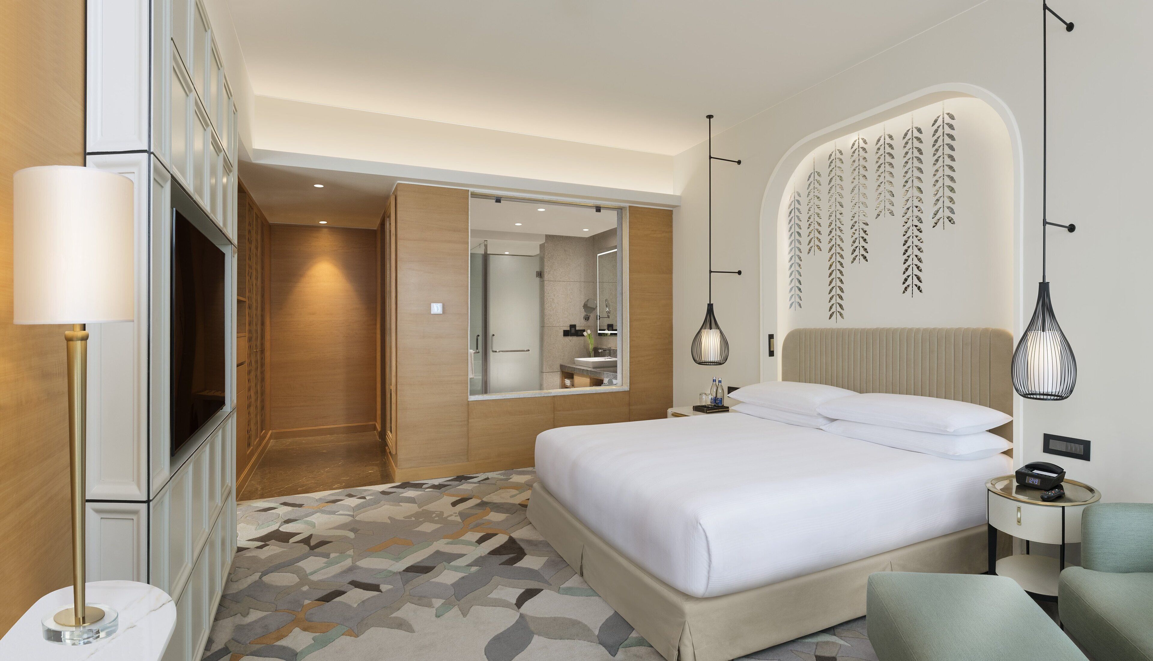 Ein Hotelzimmer mit einem großen Bett, einem Nachttisch mit Lampe, einem wandmontierten Spiegel und einem dekorativen Blattmuster an der Wand.