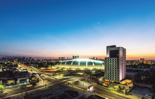 Um estádio com design moderno, cercado por um horizonte urbano com arranha-céus e uma rodovia movimentada.