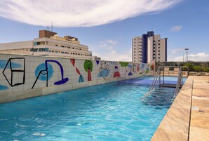 Piscina com parede revestida e murais coloridos, céu azul límpido e um edifício moderno ao fundo.