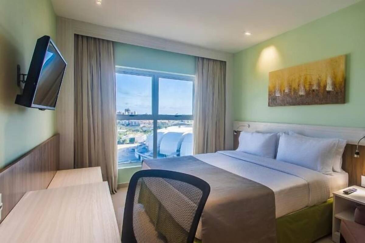 A hotel room with a bed, a desk, a chair, a TV, and a painting on the wall.