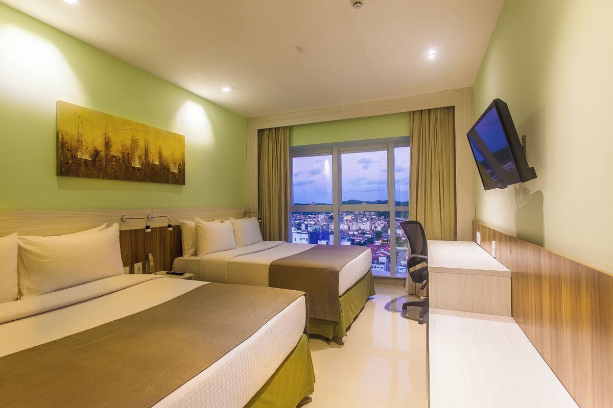 A hotel room with two beds, a desk, a chair, a television, and a large window with a city view.