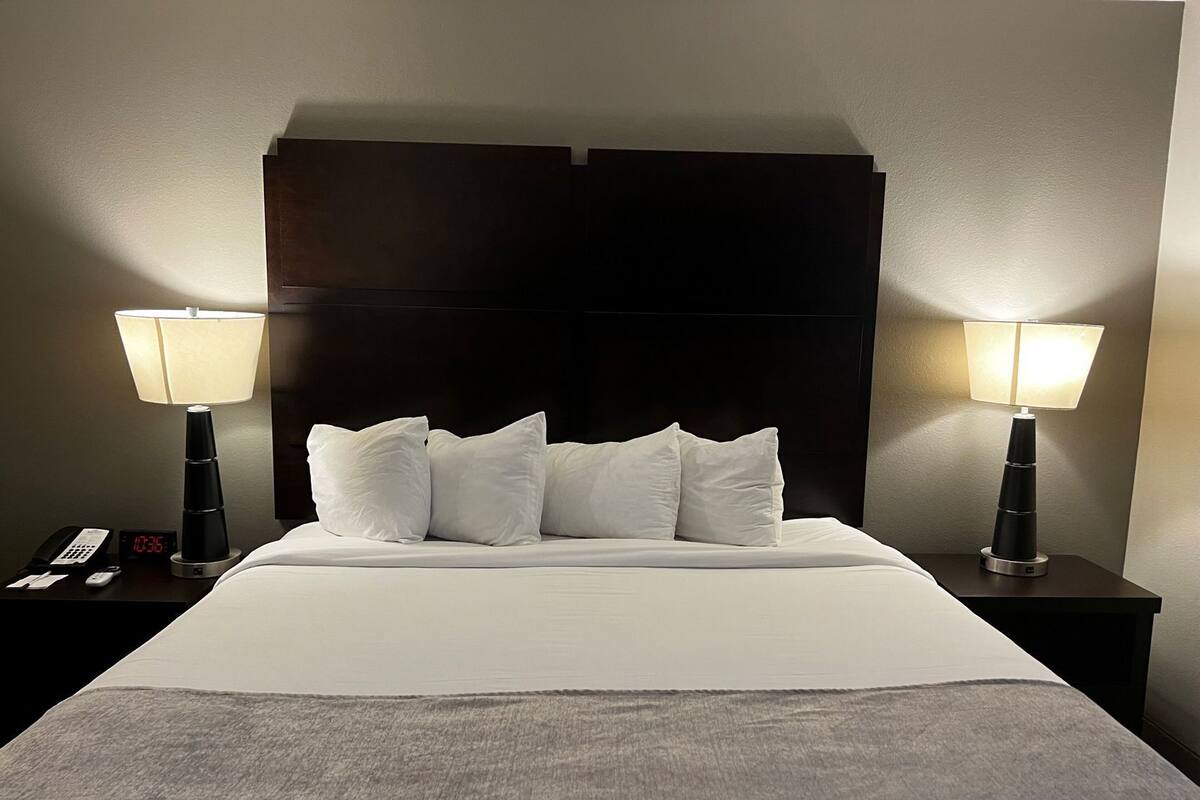 Una habitación de hotel con una cama grande, dos mesitas de noche con lámparas, una mesita con teléfono y reloj, y un cabecero oscuro.