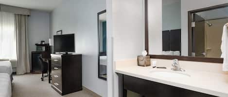 A hotel room with a bed, a desk with a computer, a television, a large mirror, and a bathroom with a sink and shower.