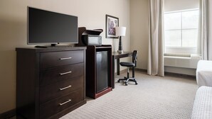 Habitación de hotel con cómoda de madera oscura, televisor de pantalla plana, microondas, escritorio con silla, un cuadro en la pared y un ventanal con cortinas.