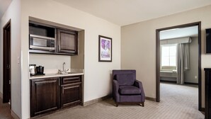 Una habitación de hotel con kitchenette, un sillón púrpura, un cuadro enmarcado y vista al pasillo.