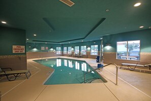 Ein Innenpool mit einem Schild 'POOL EMERGENCY SHUT OFF', Liegestühlen am Pool und Blick auf den Parkplatz durch große Fenster.