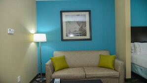 A hotel room with a beige sofa, two green pillows, a framed picture on the wall, a floor lamp, and a bed with white bedding.