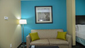 Habitación de hotel con un sofá color beige, dos almohadas verdes, un cuadro enmarcado en la pared, una lámpara de pie y una cama con ropa de cama blanca.