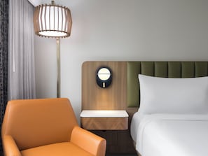 Quarto de hotel com cama, mesa de cabeceira, abajur e uma poltrona laranja.