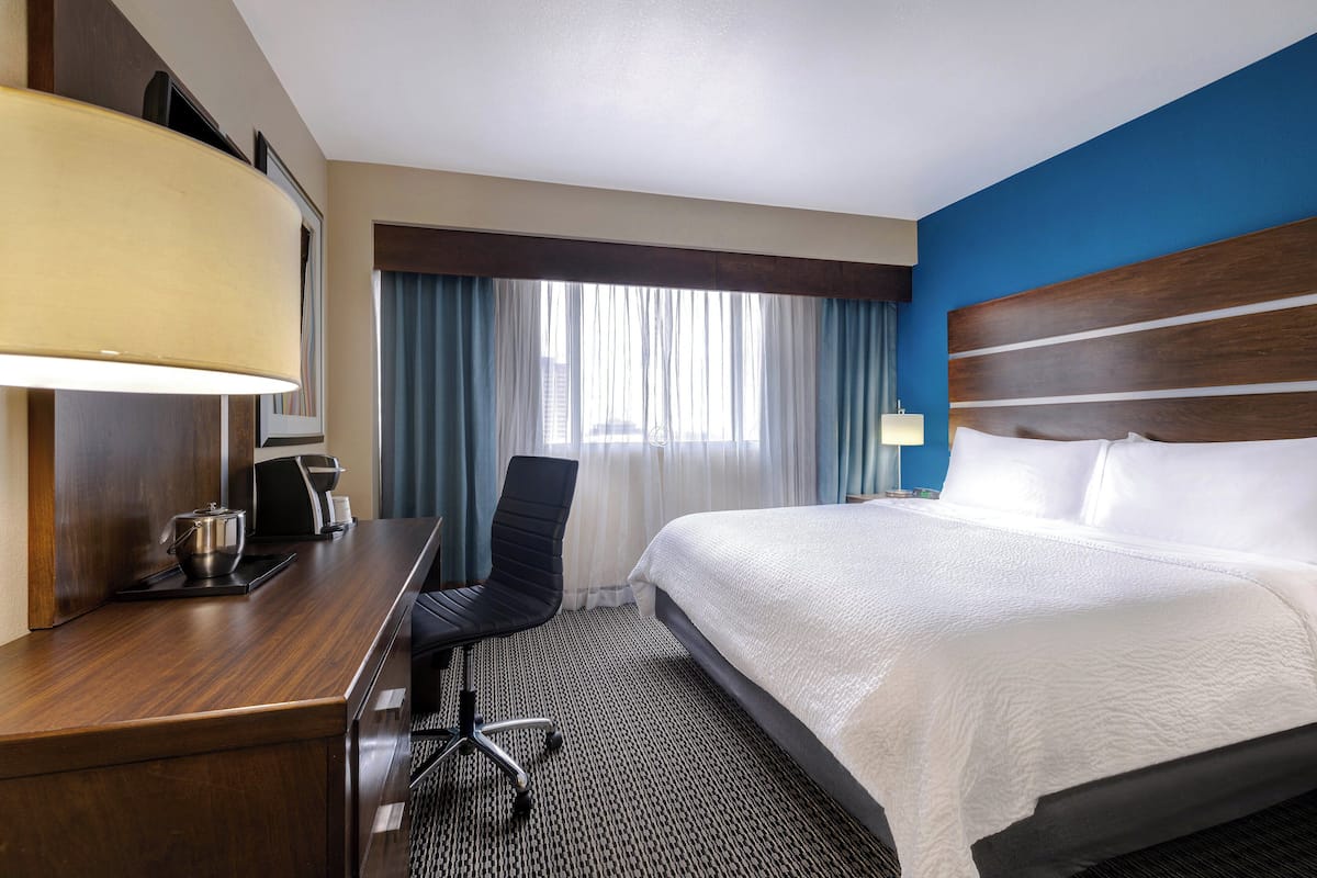 A hotel room with a bed, a desk with a lamp, a chair, and a view of the city through large windows.