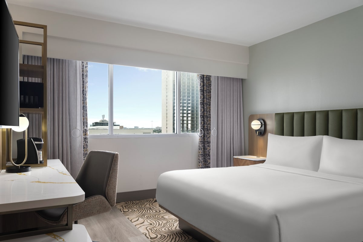 A hotel room with a bed, desk, chair, and a view of the cityscape.
