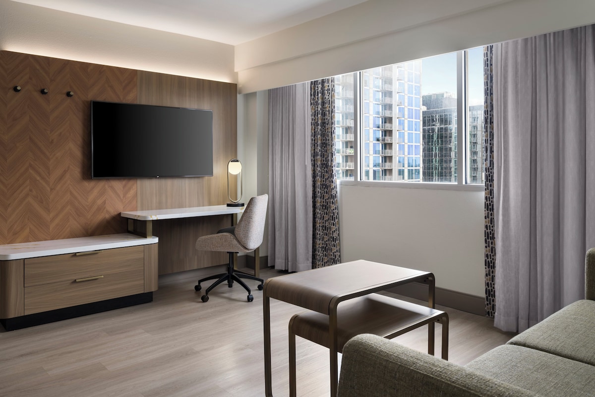 A hotel room with a flat-screen TV, a desk, a chair, a sofa, and a large window with curtains.