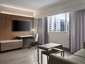 A hotel room with a flat-screen TV, a desk, a chair, a sofa, and a large window with curtains.