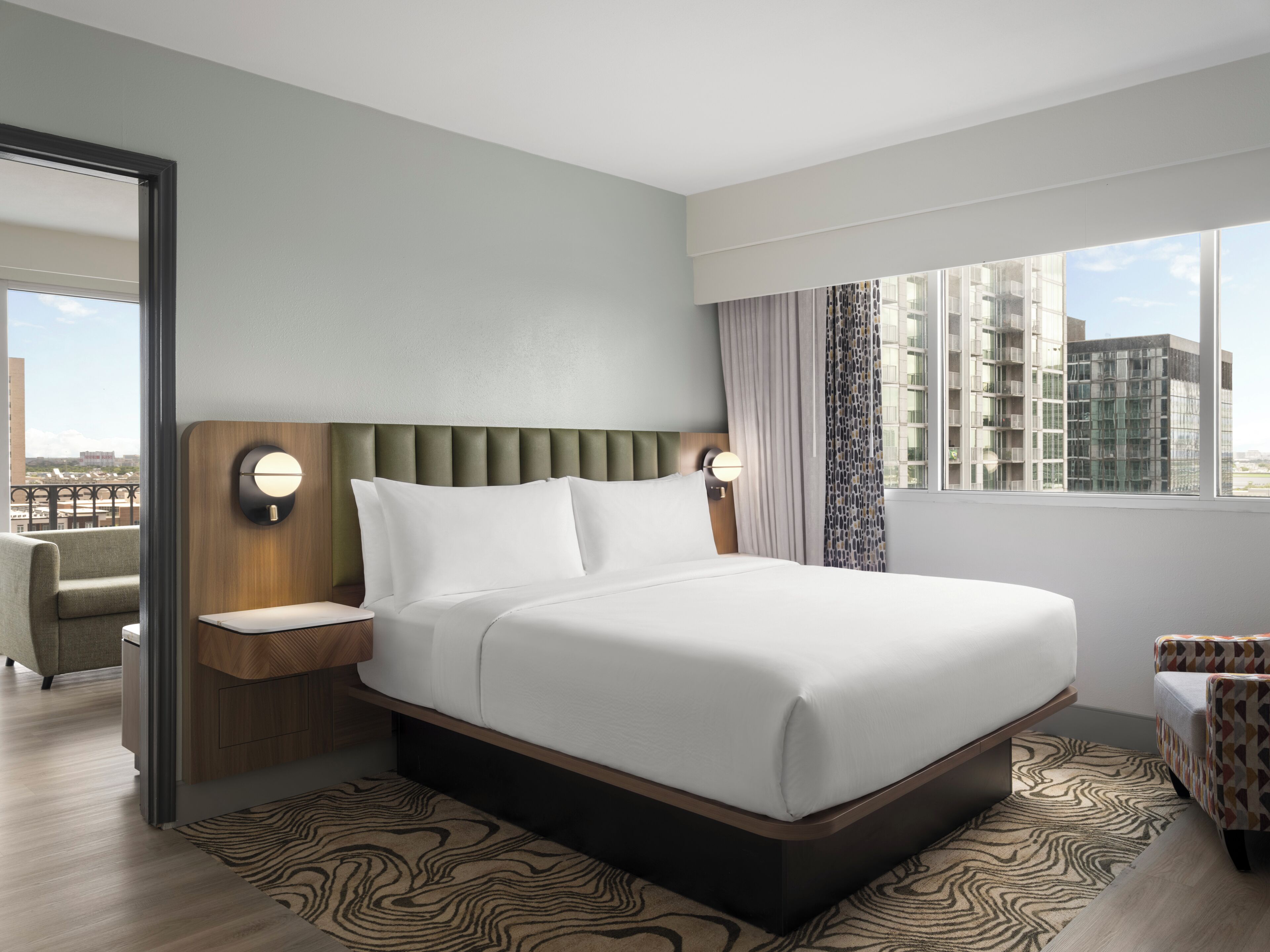 A modern hotel room with a large bed, two bedside tables, a chair, a view of the city, and a patterned rug.