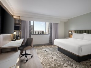 Habitación de hotel con cama, escritorio, silla y un gran ventanal con vista a edificios.