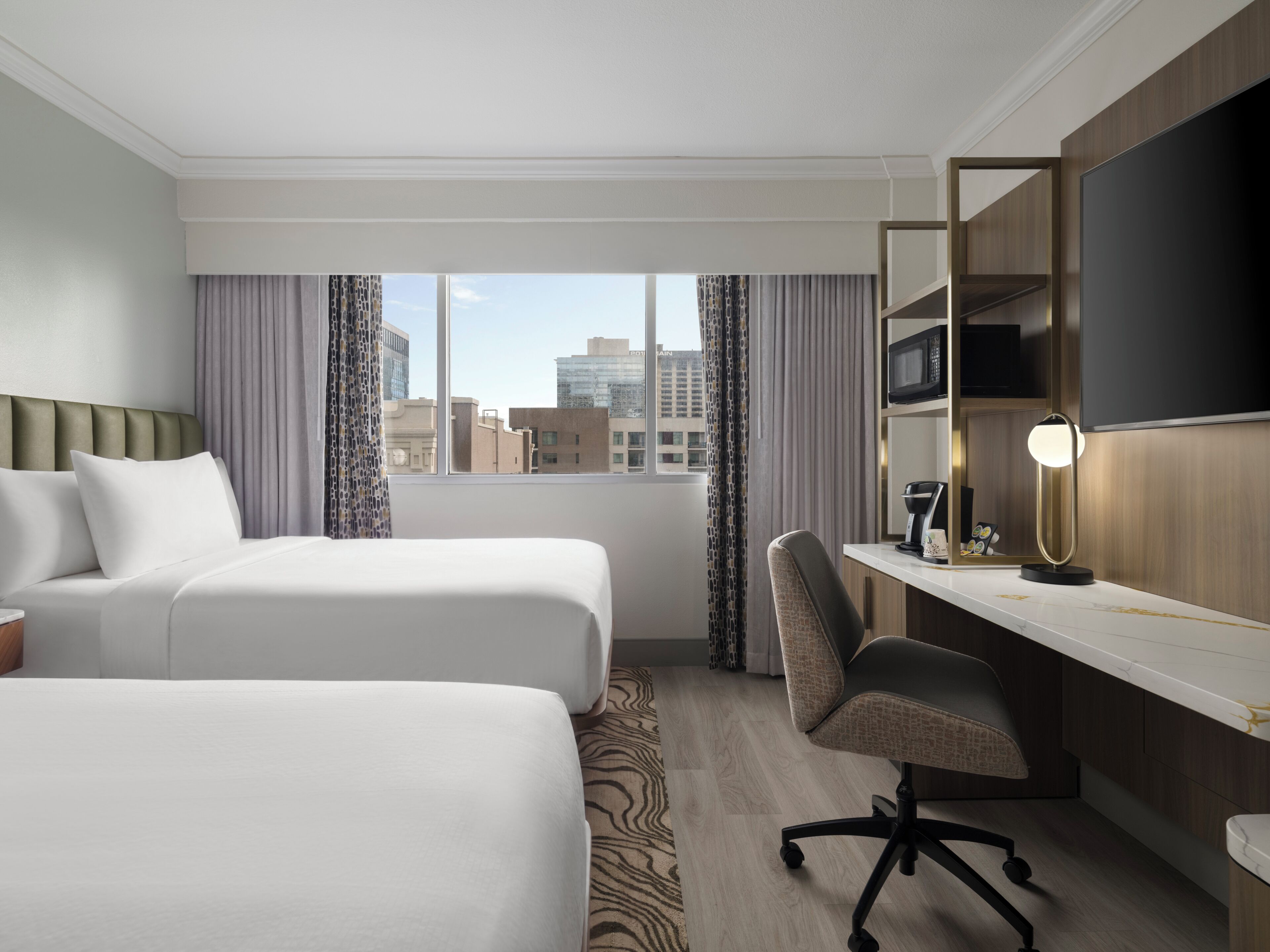 A hotel room with two beds, a desk with a chair, a flat-screen TV, and a window with curtains.