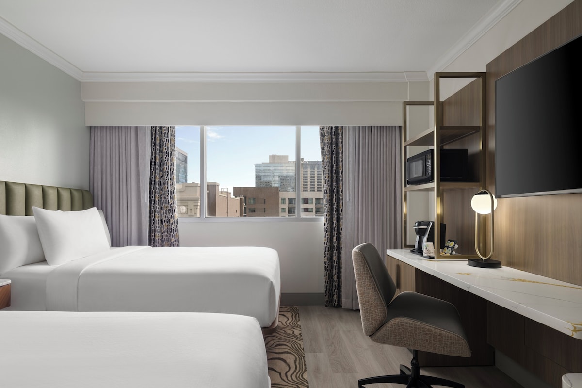 A hotel room with two beds, a desk with a chair, a flat-screen TV, and a window with curtains.