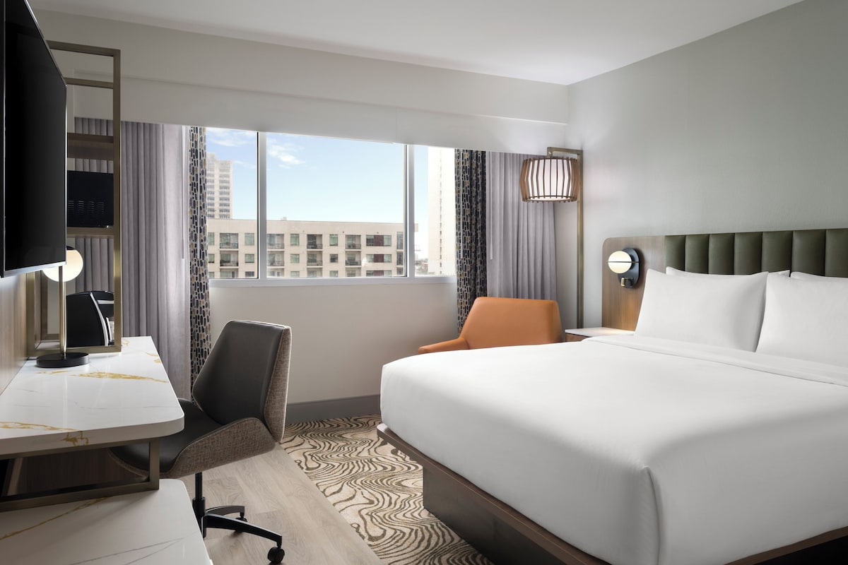 A hotel room with a bed, a desk, a chair, a TV, and a window with a city view.