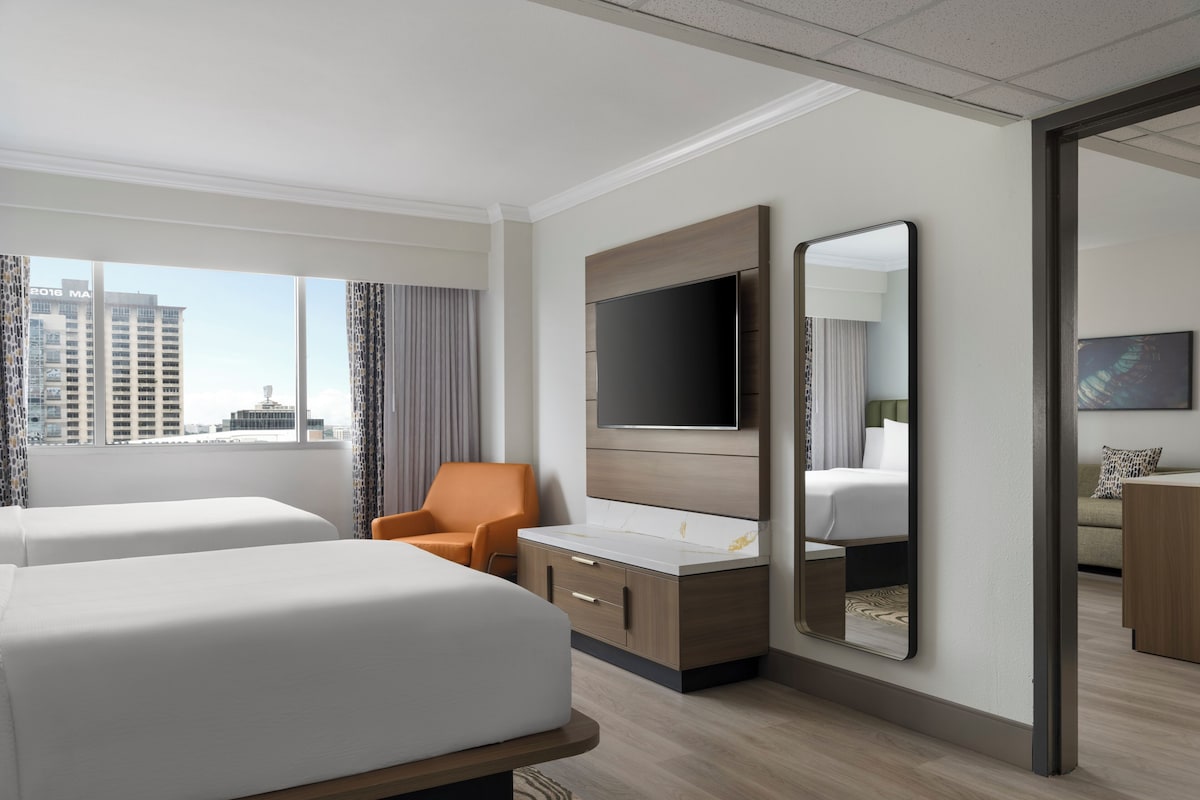 A hotel room with two beds, a TV, a desk, and a large window with a city view.