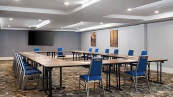 Un salón de conferencias con una mesa larga, sillas azules, una televisión de pantalla plana y alfombra estampada.