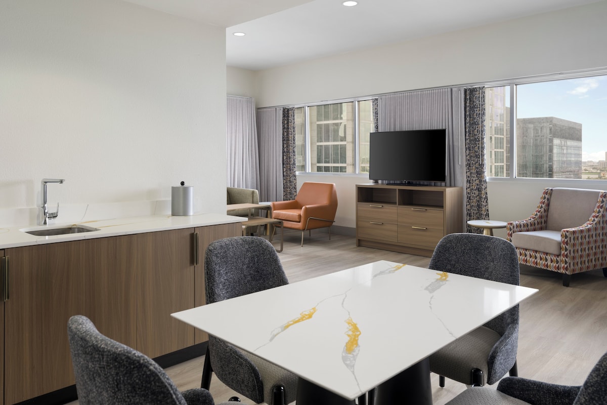 A modern hotel room with a dining area, a sofa, a TV, and a large window.