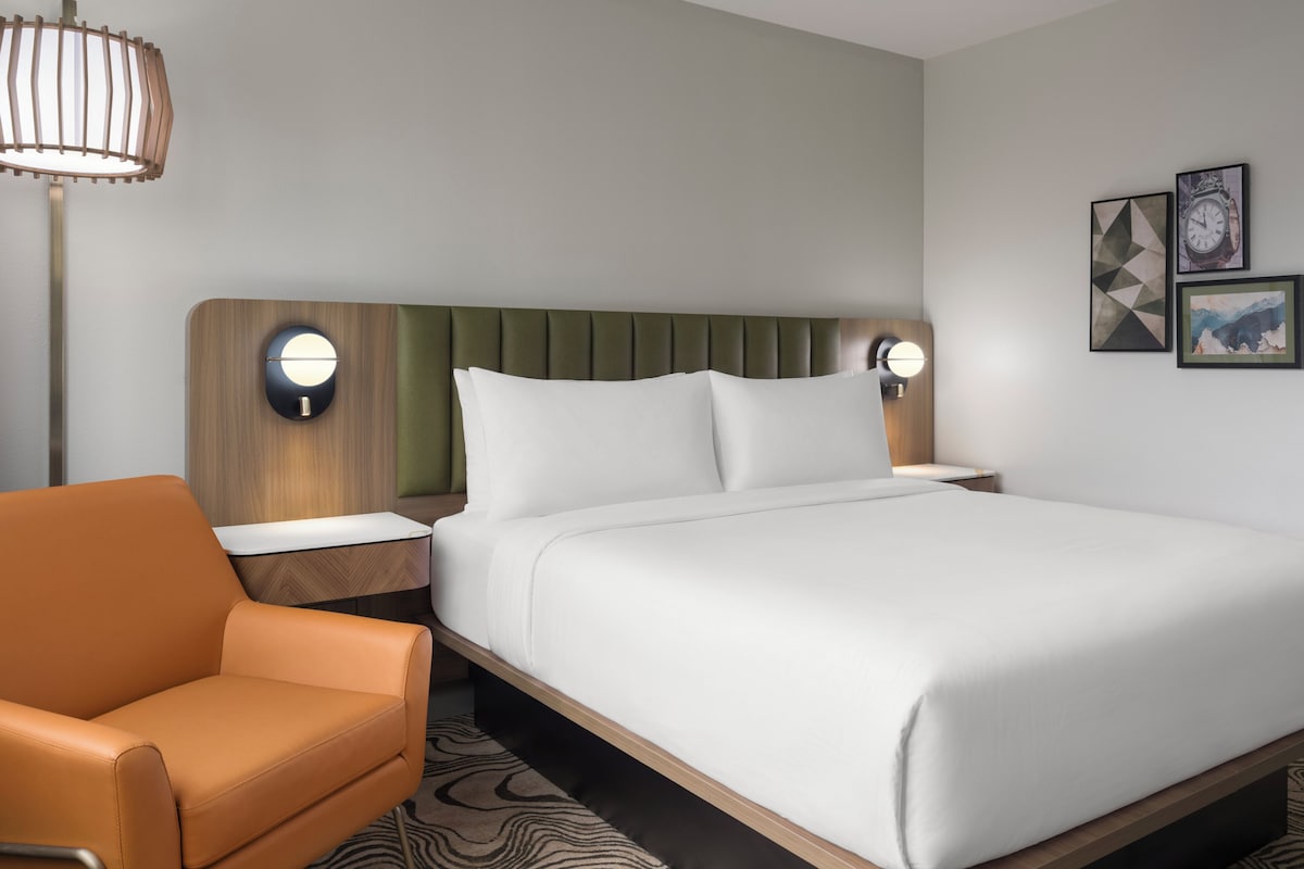 A hotel room with a bed, a nightstand, a lamp, a chair, and wall art.
