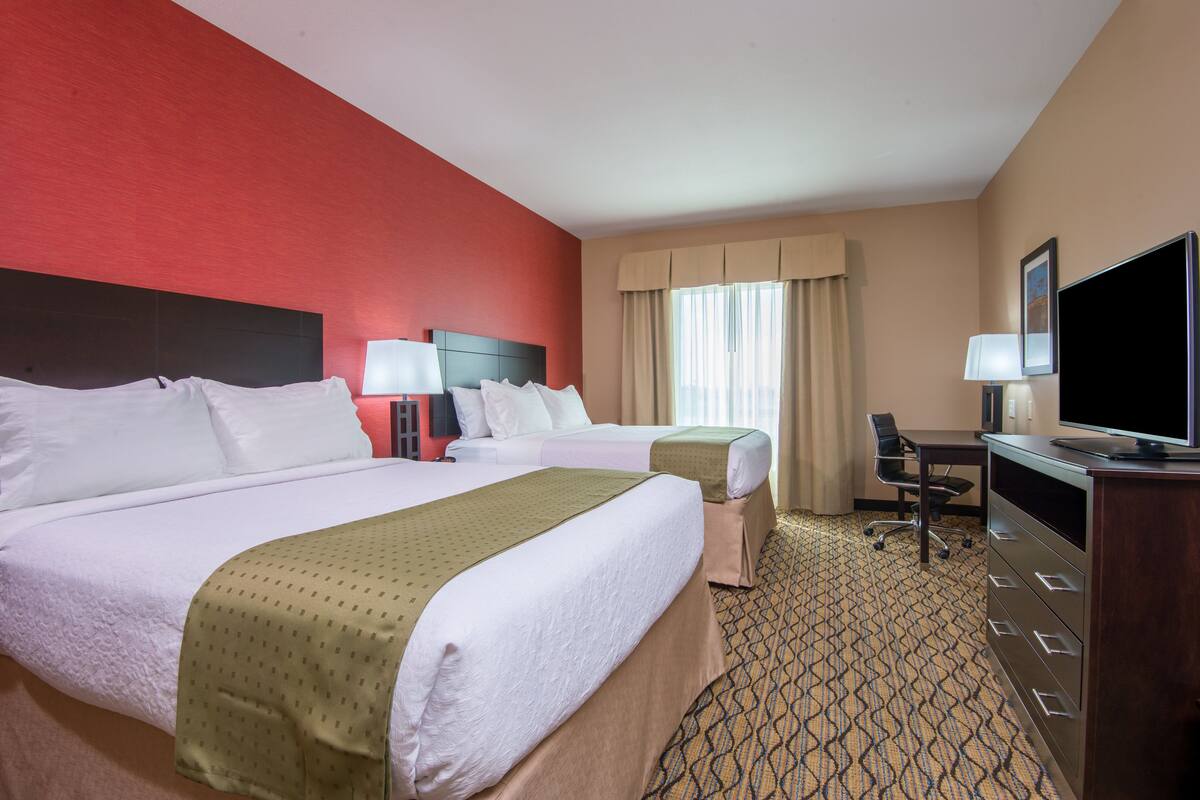 A hotel room with two beds, a desk, a TV, and a window with curtains.