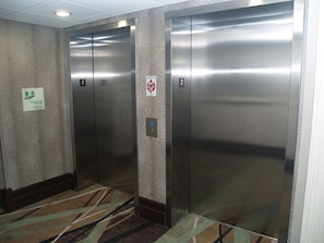 Dos puertas de ascensor en un pasillo con alfombra estampada y un cartel que indica el uso de las escaleras en caso de incendio.