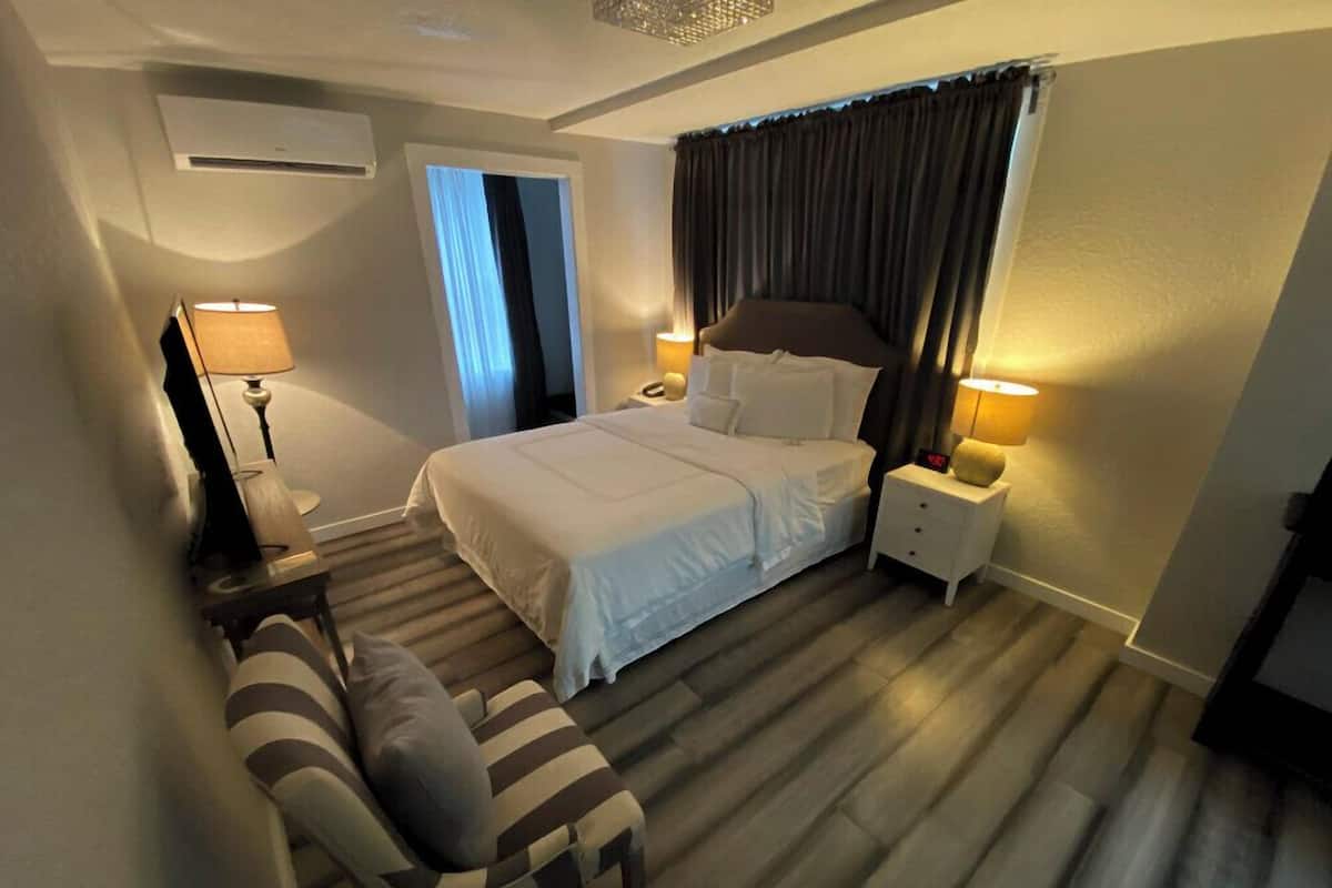 A hotel room with a bed, bedside lamps, a chair, a window with curtains, and an air conditioning unit.