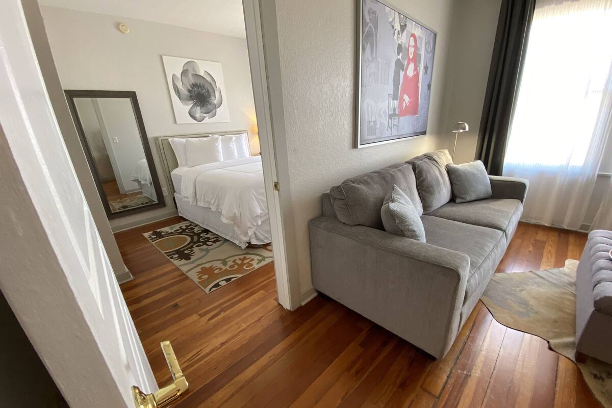 A room with a grey sofa, a bed with white bedding, a large mirror, and a framed artwork on the wall.