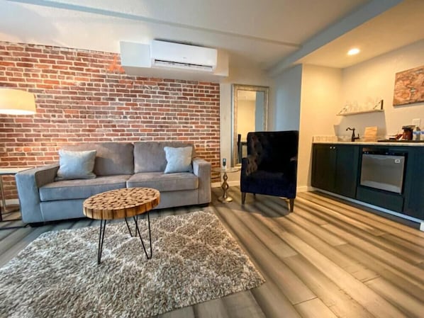 A living room with a grey sofa, a round wooden coffee table, and a black armchair. A brick wall and an air conditioning unit are visible.