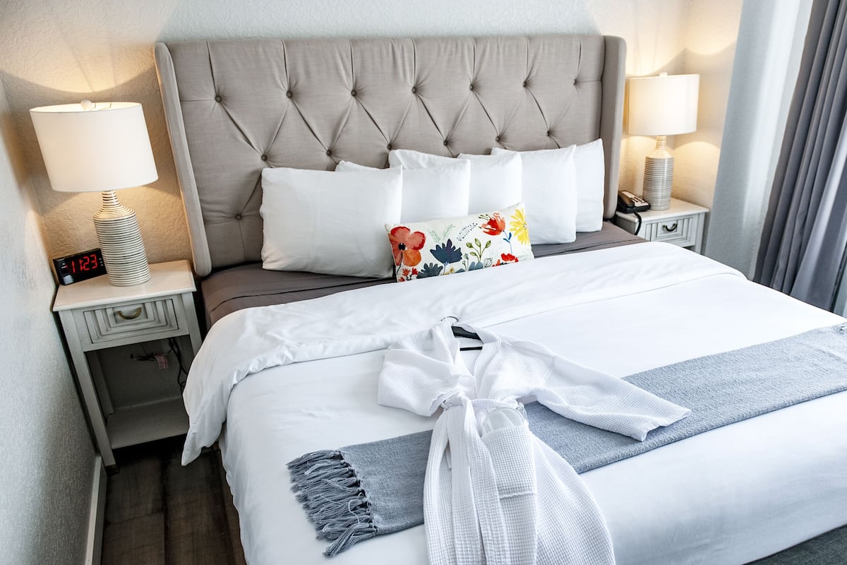 A neatly made bed with a white duvet, a grey throw blanket, and a white bathrobe tied on the bed. There are two bedside tables with lamps, a clock, and a phone. The headboard is tufted and grey.