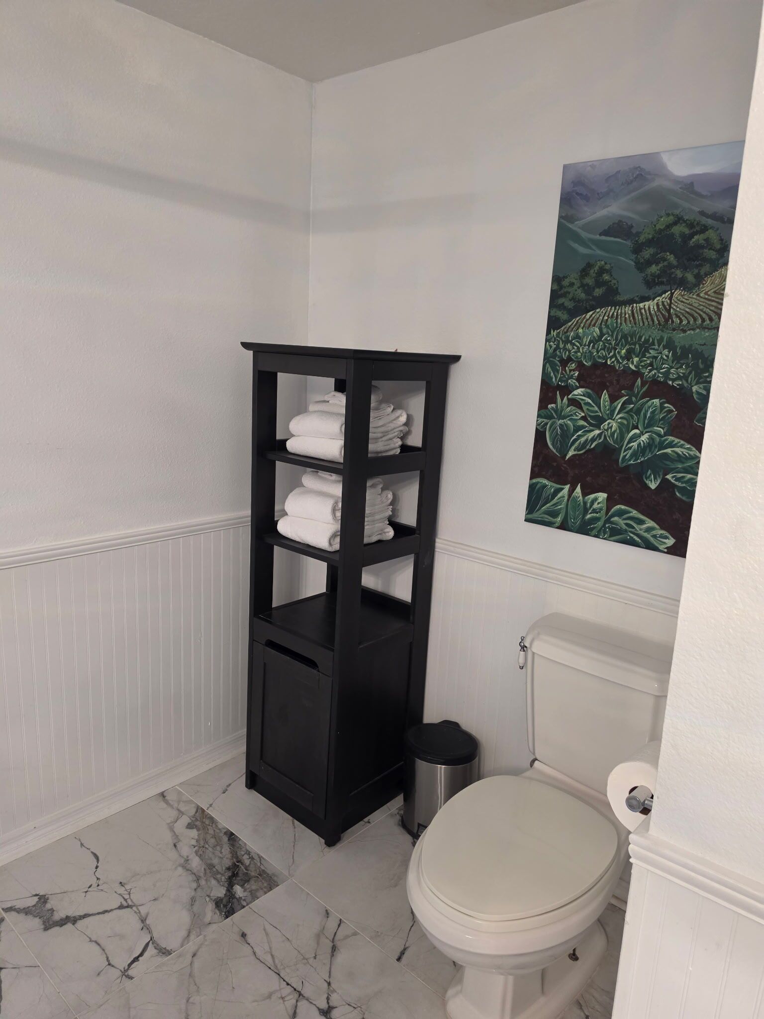 A bathroom with a marble floor, a white toilet, a black towel rack, and a painting on the wall.
