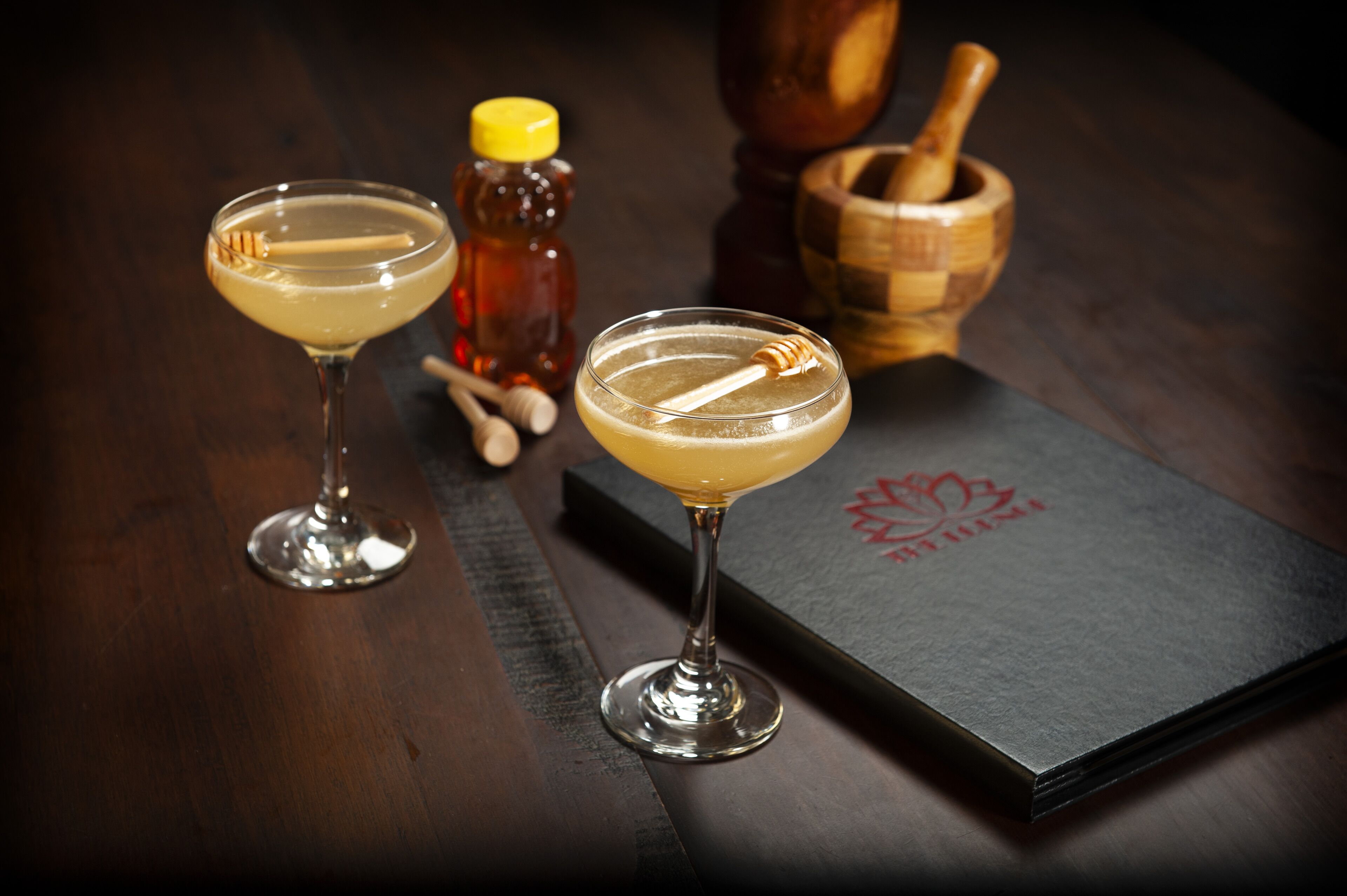 Two cocktails on a dark wooden table with a honey bottle and a wooden mortar and pestle.