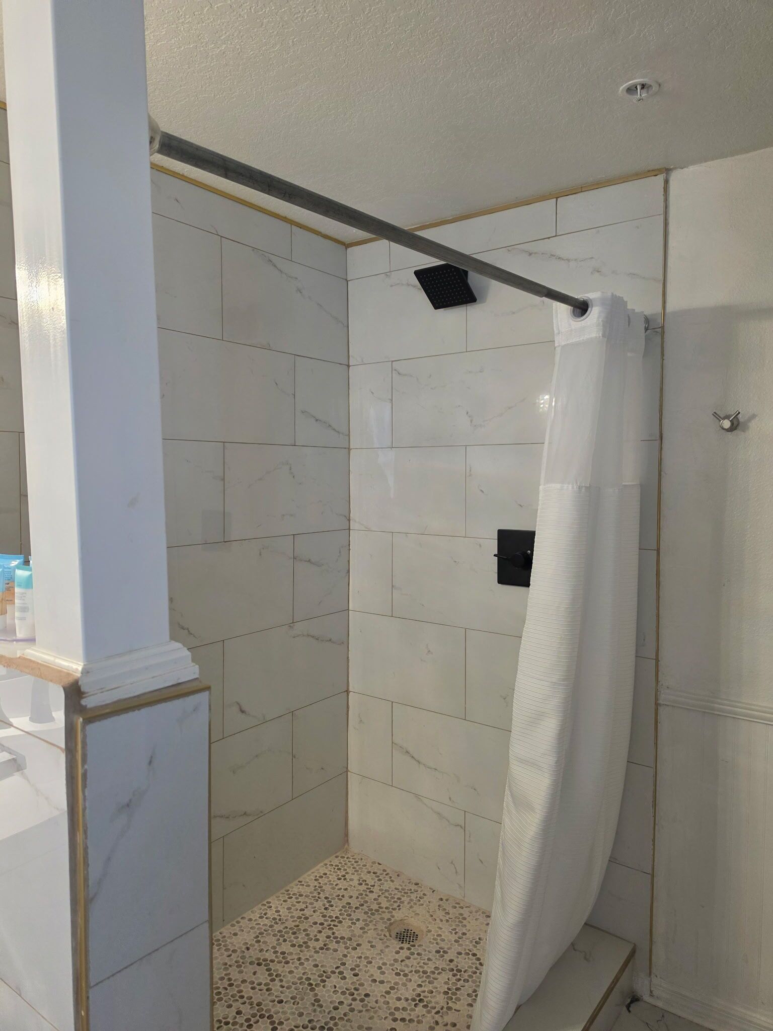 Une douche avec un rideau blanc, des murs carrelés et une barre métallique pour suspendre des objets.