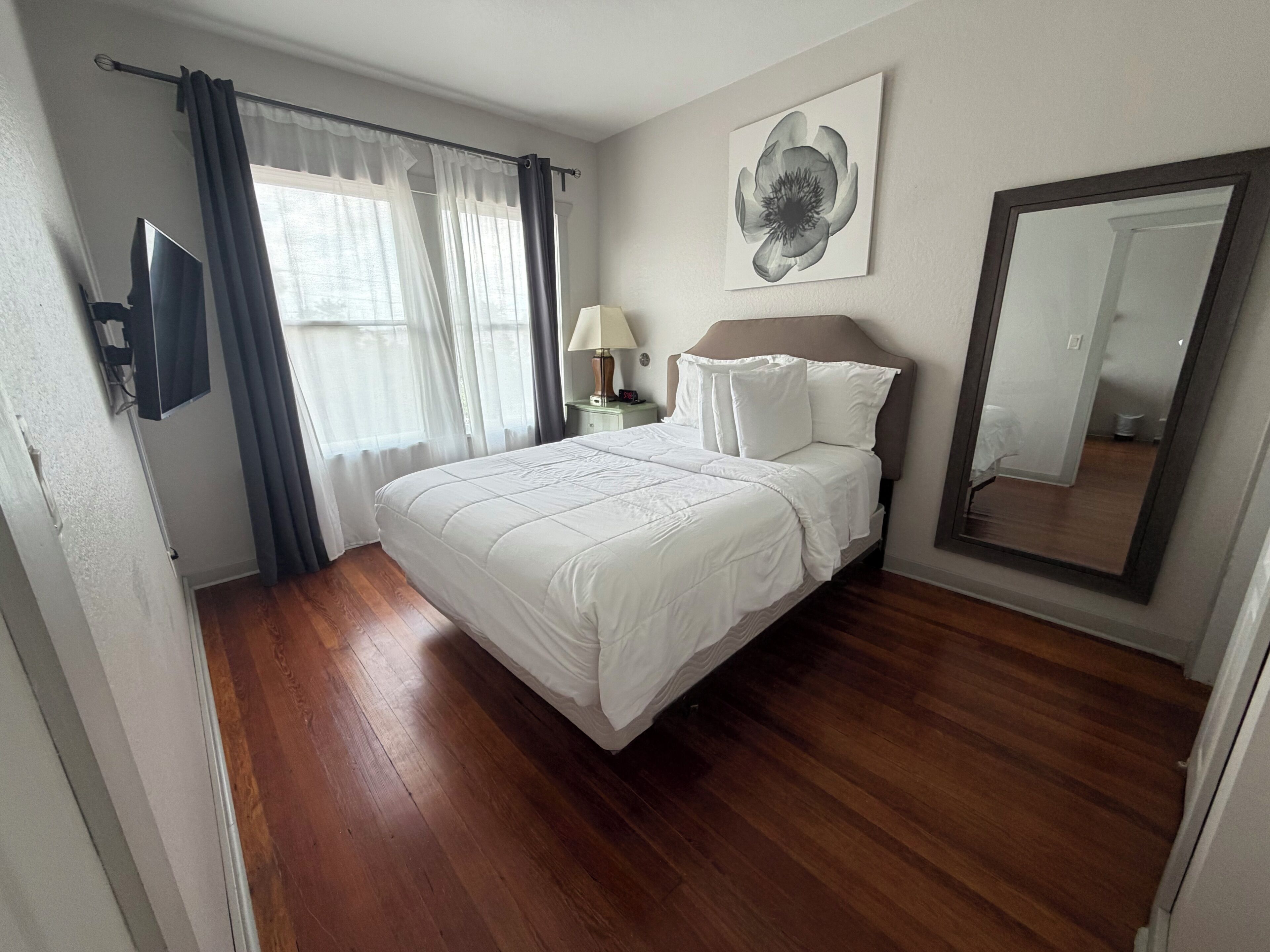 Une chambre à coucher comprenant un lit, une table de chevet, une lampe, une fenêtre avec des rideaux, une œuvre d’art accrochée au mur et un miroir.