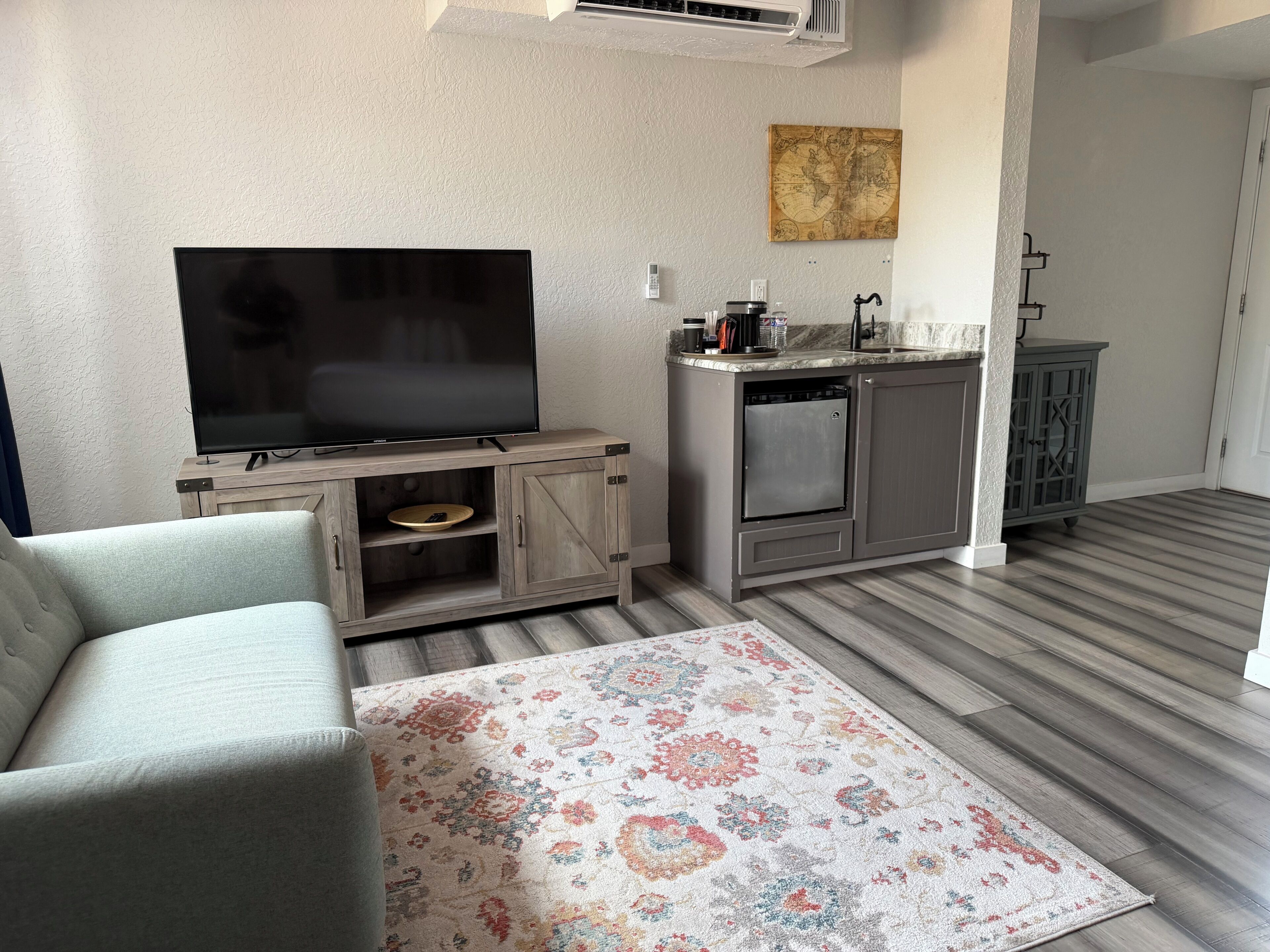 A living room with a flat-screen TV, a gray sofa, a patterned rug, and a kitchen area with a sink and cabinets.