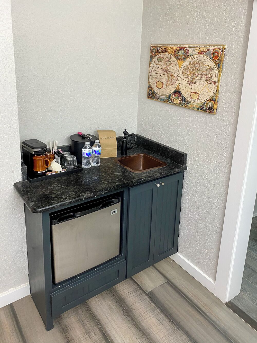 A compact kitchen with dark cabinets, a stainless steel dishwasher, a black countertop, and a decorative world map on the wall.