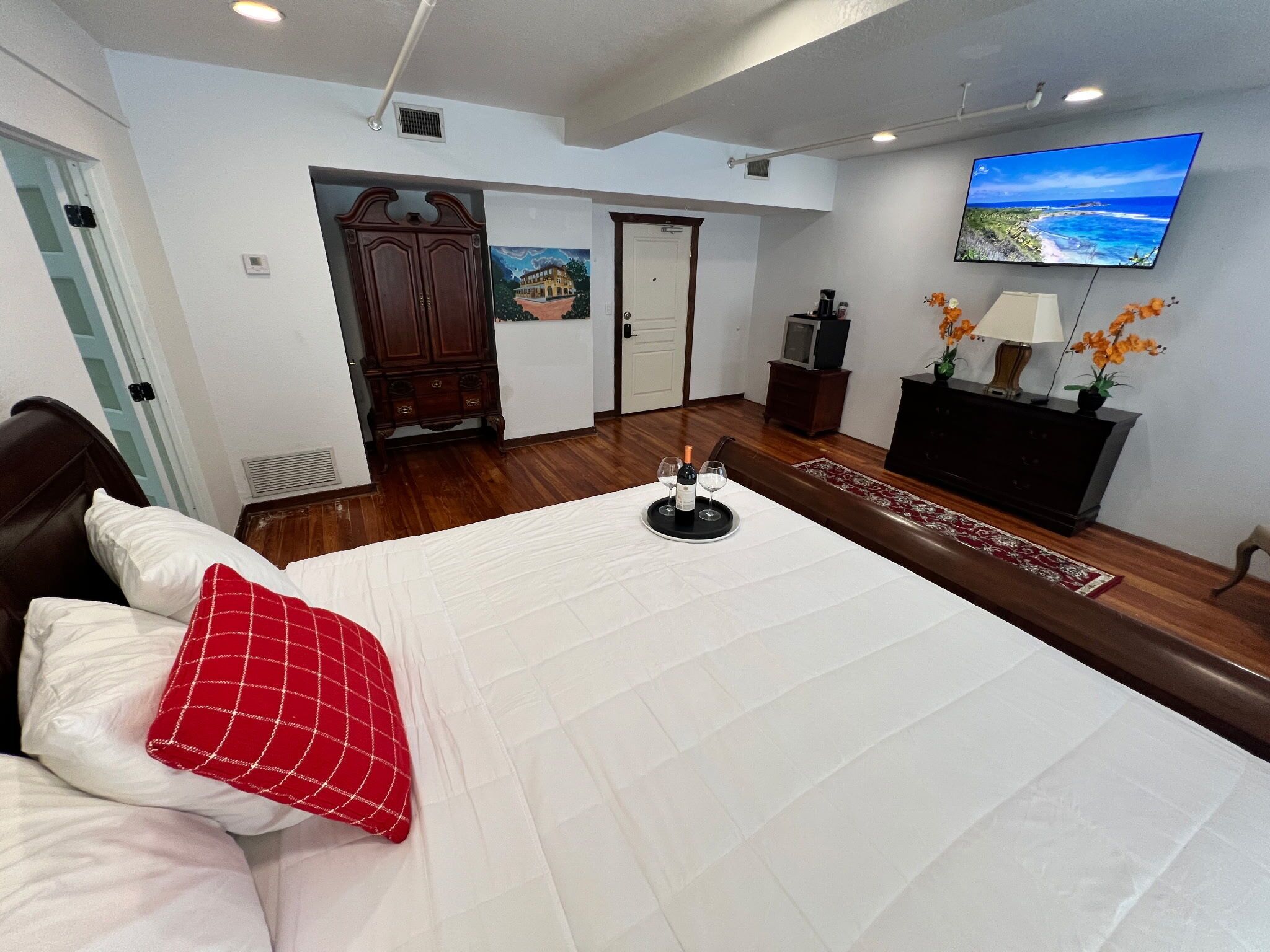 A bedroom with a bed, a TV mounted on the wall, a wooden dresser, and a painting of a building.