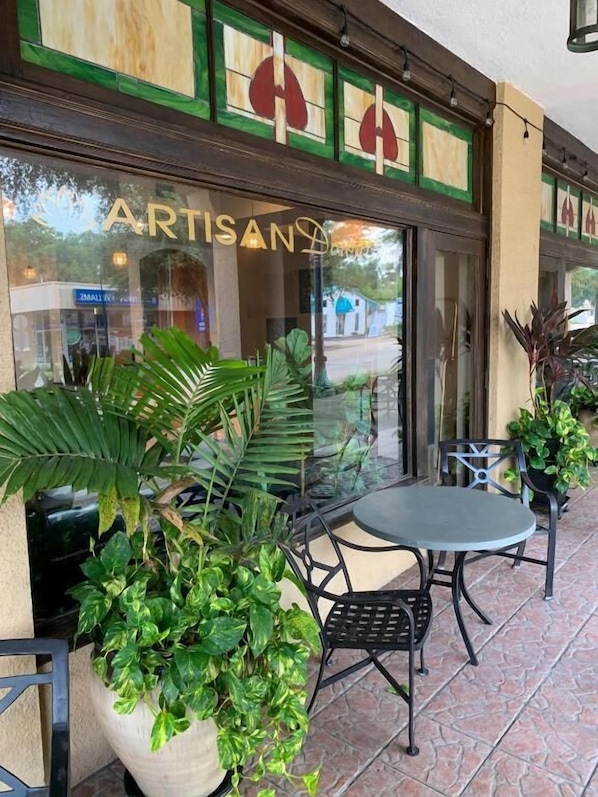 An outdoor seating area with tables and chairs, potted plants, and a storefront with a sign reading "ARTISAN."