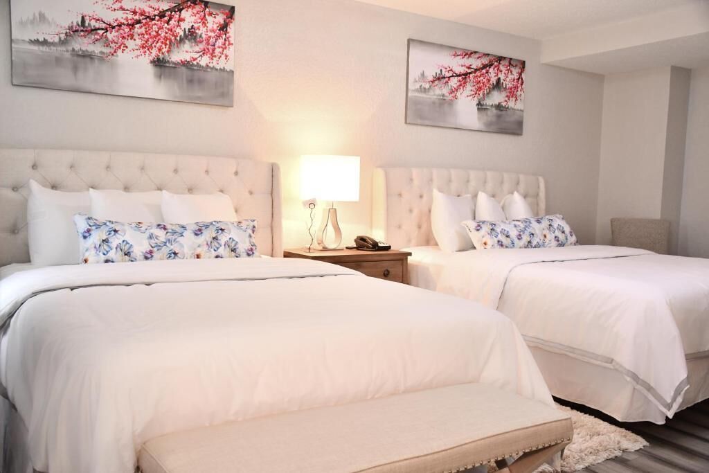 Habitación de hotel con dos camas, cada una con ropa de cama blanca y almohadas estampadas con motivos florales. Hay dos mesitas de noche con lámparas y un teléfono. La habitación tiene una paleta de colores neutra y cuadros decorativos en las paredes.