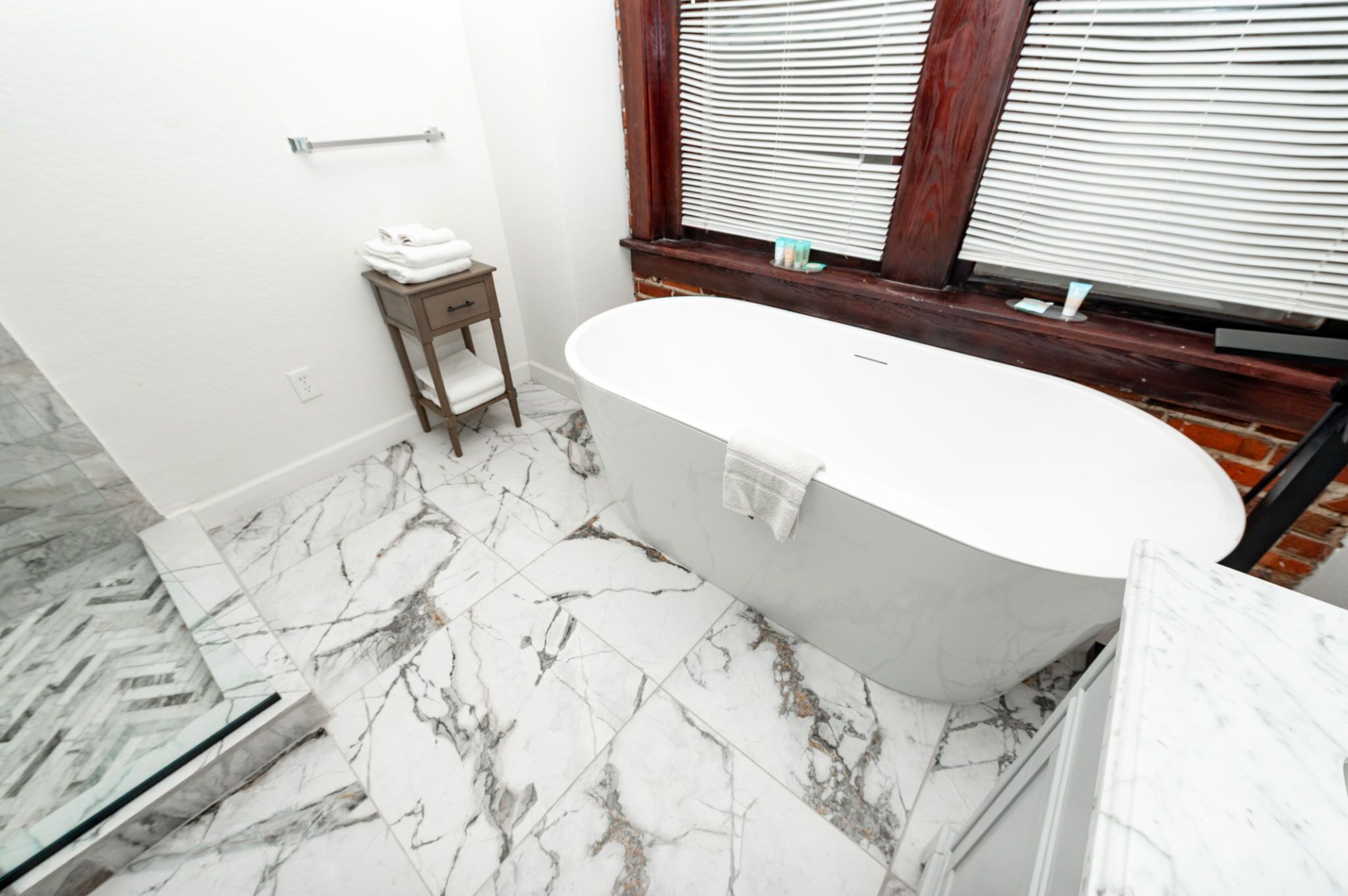 A bathroom with a freestanding bathtub, marble flooring, a wooden side table with towels, and a window with blinds.
