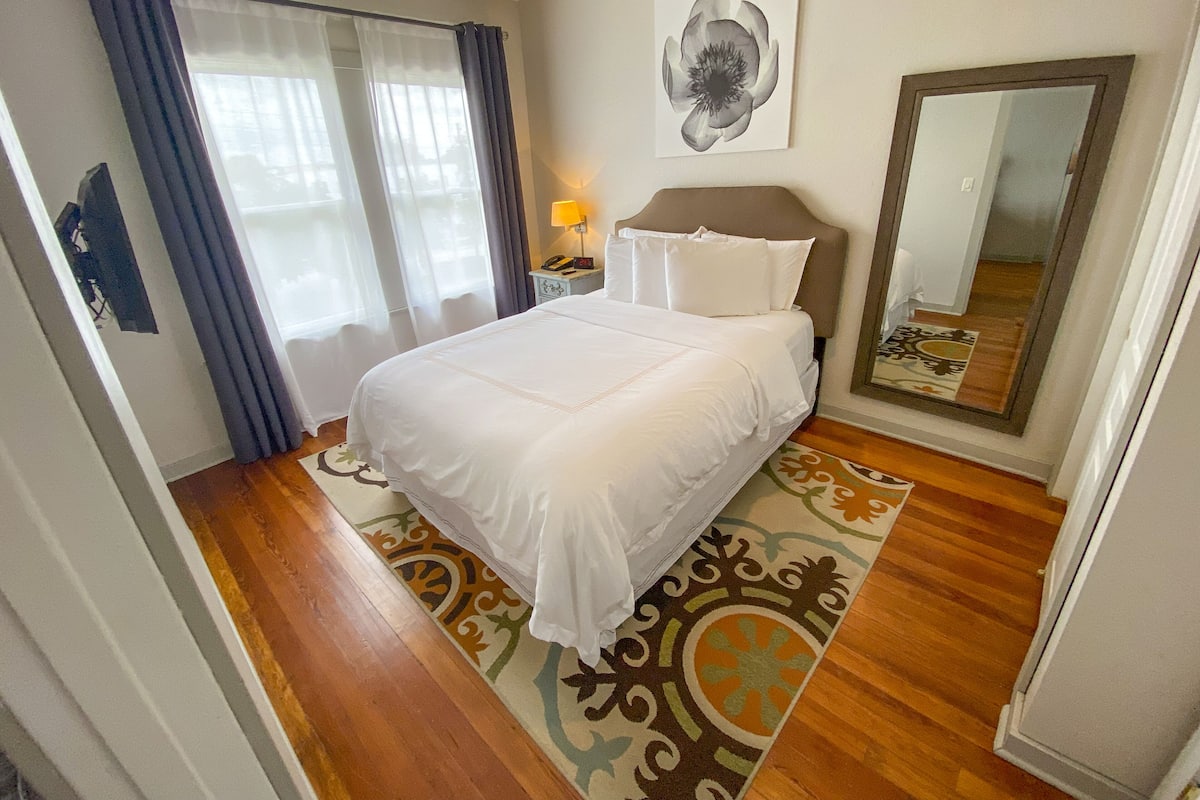 A neatly made bed with white linens, a bedside lamp, a large mirror, and a floral-patterned rug.